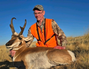 A seasoned hunter in a bright orange vest poses with a pronghorn antelope lying on the prairie grass, showcasing its elegant horns and striking markings.